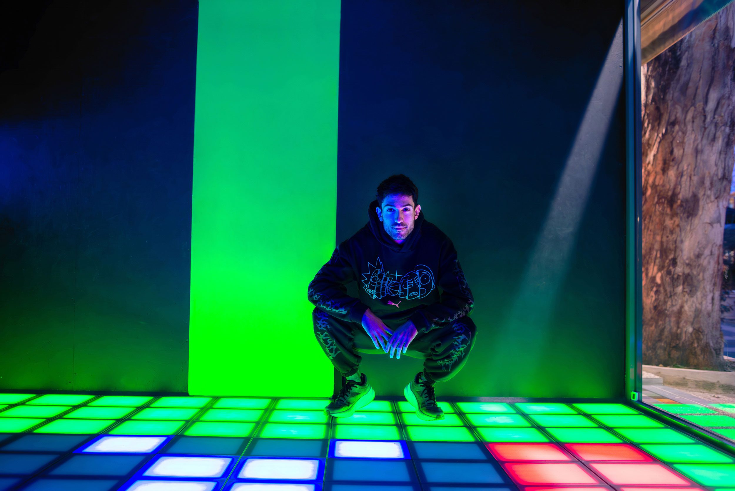 Ο Μίλτος Τεντόγλου κάθεται χαμηλά και το πάτωμα από κάτω του εκπέμπει διάφορα χρώματα.